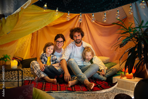 Amazing family time spending two cute and pretty boys and their parents sitting on the floor inside the handmade tent in the kids room eating popcorn and watching a movie on the laptop