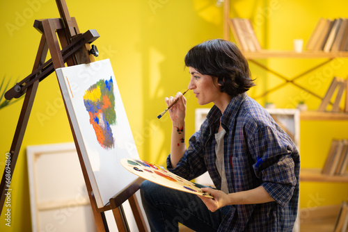 A painter is holding a very large palette with his hands and squeezing paint onto it so he could continue to paint on his canvas