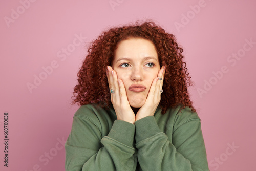 Obraz na plátne Studio portrait of pretty bored funny ginger head millennial girl with cute curls touching her cheeks and pouting lips looking aside isolated on pink background