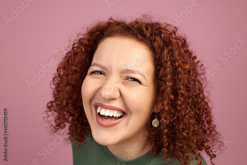 Studio headshot of cheerful laughing woman with red curls, demonstrating white teeth, isolated against pink background. Overjoyed young cute female having fun. Dental health, malocclusion photo
