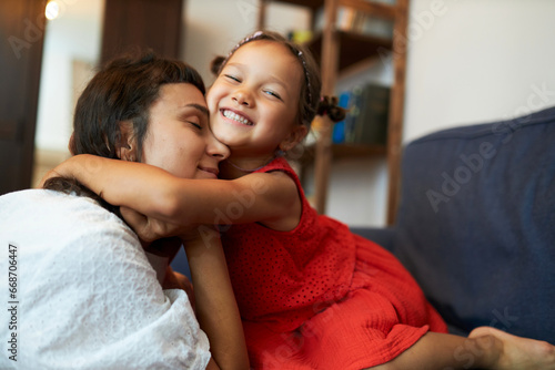 Side view of cute happy smiling little girl in red dress hugging closely her mother on sofa, demonstrating affection, woman cuddling with kid, showing unconditional love for her child