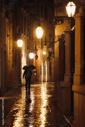 Person walking in the rain at night. Man with an umbrella. Old historical street with warm lamps. 