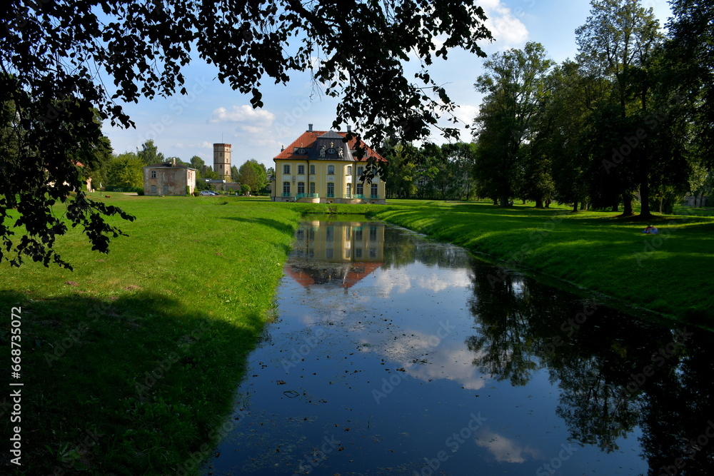 Obraz premium A view of a channel or lake overgrown with moss and vines, with forests and moors on both sides combined with small concrete bridges, walkways, orchards, and a single manor in the middle