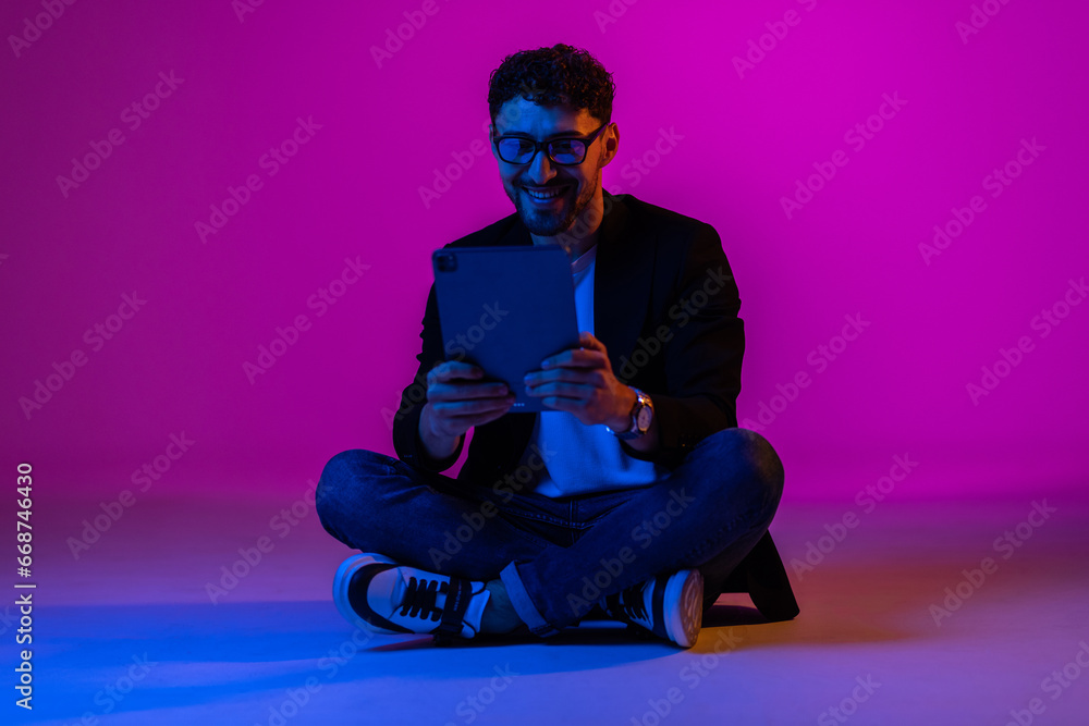 Young man wearing business suit and eyeglasses using digital tablet on gradient blue purple background in neon light. Young man wearing business suit and eyeglasses using digital tablet on gradient blue purple background in neon light.