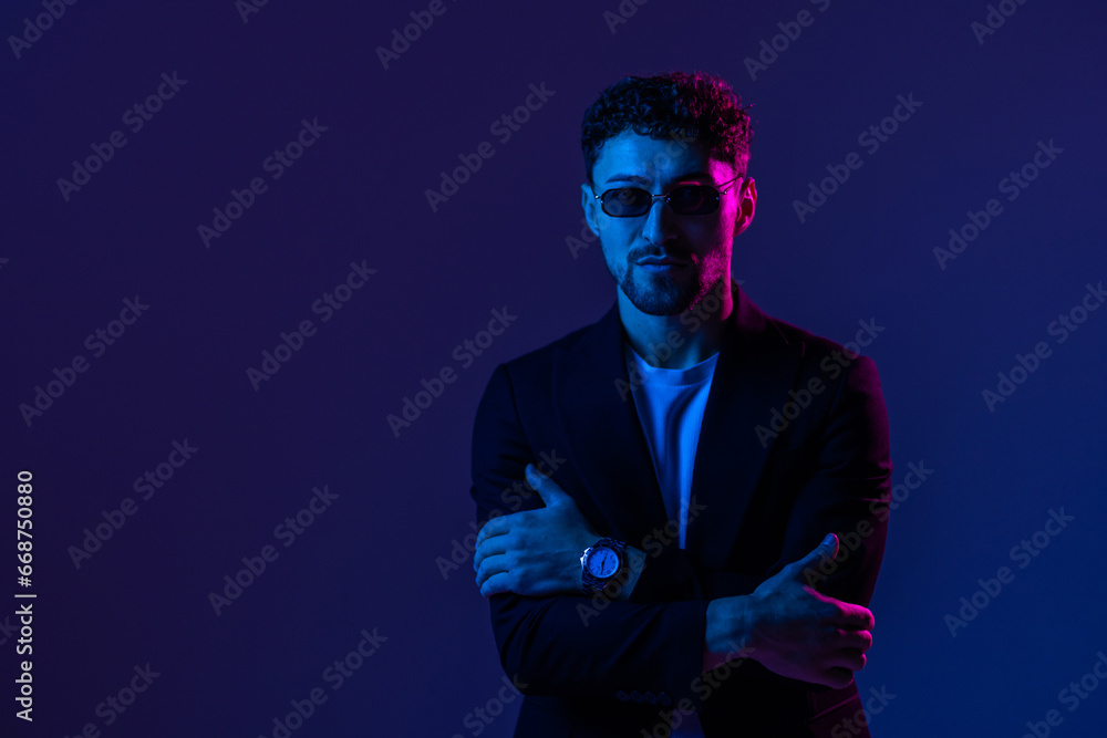Young man with beard wearing sun glasses and a leather jacket between red and blue neon lights