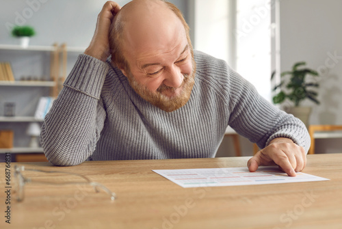 Senior man with dementia gets confused and scratches head when he looks at calendar on table. Old man with Alzheimer's disease can't remember important dates, events, birthdays, hospital appointments photo