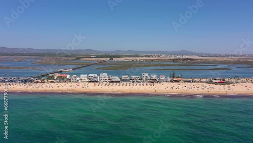 Aerial view of the ocean coast and beach. Boats on water and tourists relaxing on the beach as seen from a drone. Praia de Faro Algarve Portugal. Airport and city in the background.