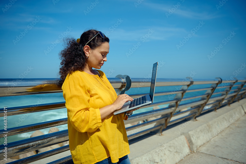 Side portrait of multitasking confident female entrepreneur working remotely on laptop, standing against sea background