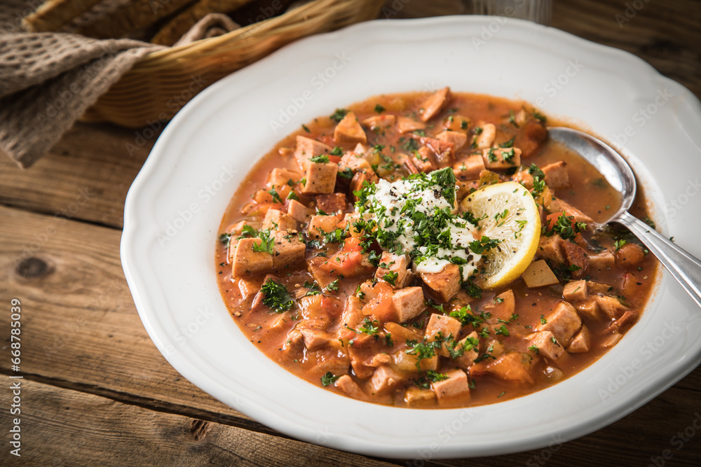 Ost Deutsche DDR GDR Soljanka Eintopf Suppe mit Jagdwurst, Saure Gurken, Letscho, Schmand, Zitrone, Toast und Petersilie auf Holzbrett mit Brotkorb, Salzstreuer auf Holz Hintergrund Ost Deutsche DDR GDR Soljanka Eintopf Suppe mit Jagdwurst, Saure Gurken, Letscho, Schmand, Zitrone, Toast und Petersilie auf Holzbrett mit Brotkorb, Salzstreuer auf Holz Hintergrund