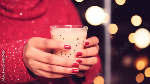 Female hands with red manicure hold a glass of coffee or other milk drink. Festive atmosphere, garland lights. Girl in a sweater with a drink, close-up