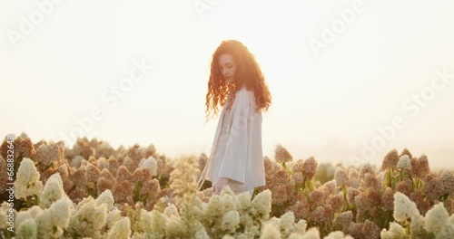 Mystical woman with red curly hair walking through flowering white shrubs.