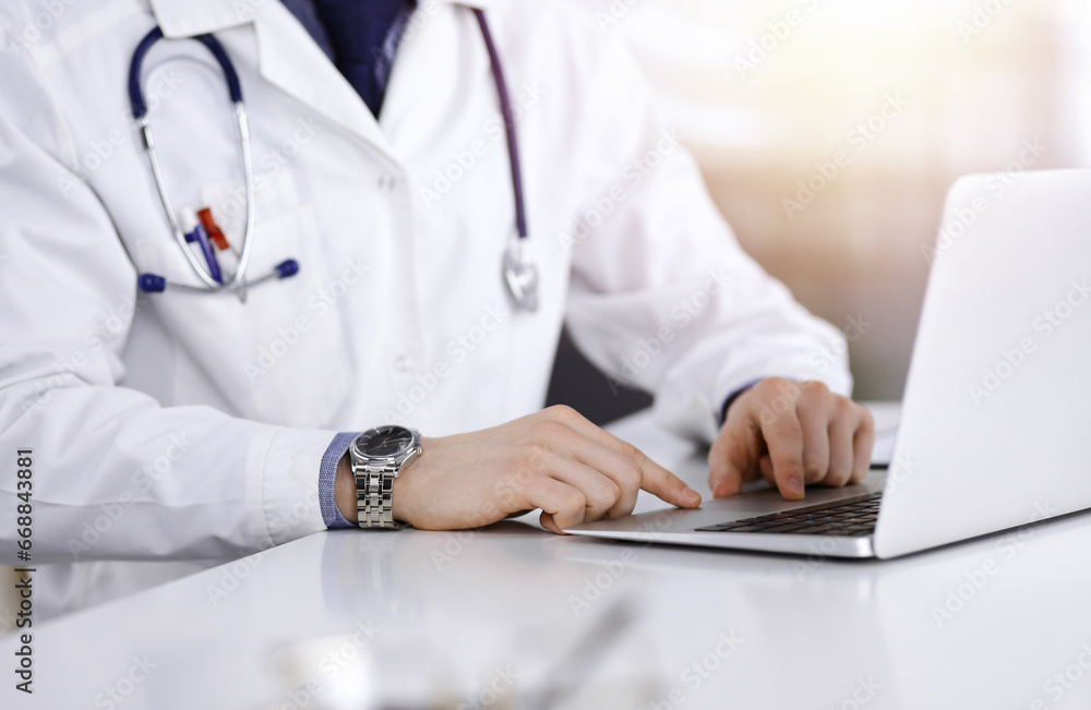 Unknown male doctor sitting and working with laptop in a darkened clinic, glare of light on the background, close-upof hands Unknown male doctor sitting and working with laptop in a darkened clinic, glare of light on the background, close-upof hands