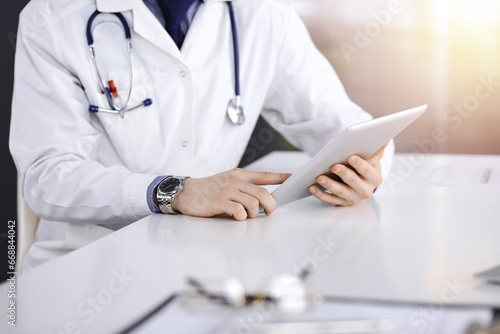 Unknown male doctor sitting and working with tablet computer iin a darkened clinic  glare of light on the background  close-up of hands