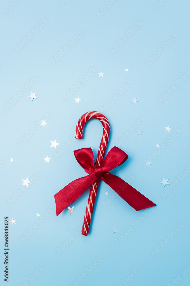 Candy cane decorated with red bow and stars confetti on blue background. Minimal Christmas concept. Selective focus, copy space Candy cane decorated with red bow and stars confetti on blue background. Minimal Christmas concept. Selective focus, copy space