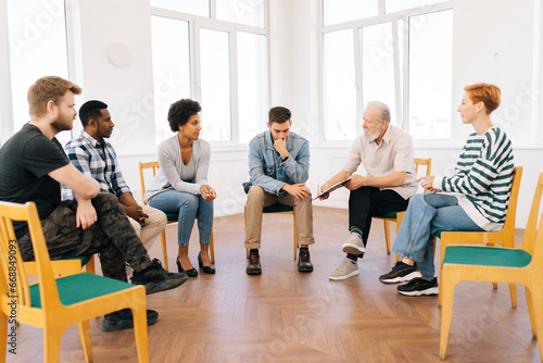 Wide shot of bearded aged professional male mentor leader counselor coach speaking at diverse business meeting with team people or patients group, during training psychotherapy session.