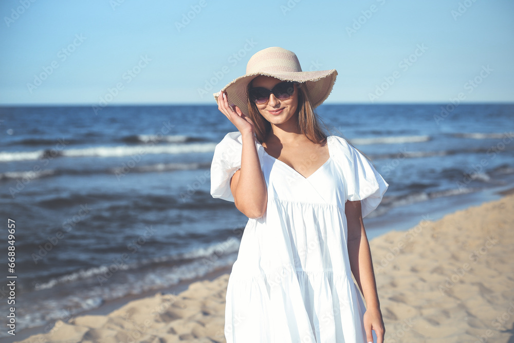 Happy smiling blonde woman is posing on the ocean beach with sunglasses and a hat