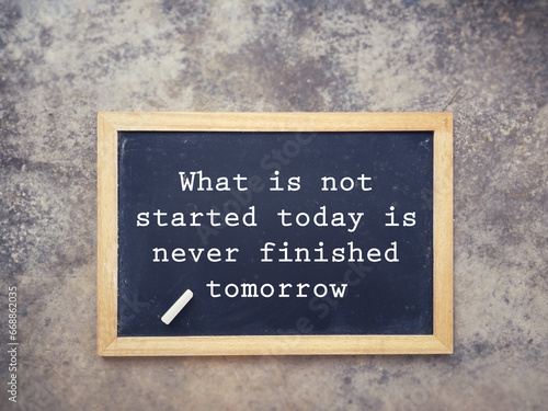 Motivational and inspirational wording. What Is Not Started Today Is Never Finished Tomorrow written on a blackboard. With blurred styled background.