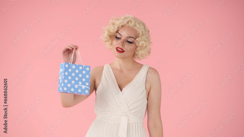 Woman holding a small blue gift bag. Woman looking like Marilyn Monroe in studio on pink background. Shopping, sale, holiday concept. Woman holding a small blue gift bag. Woman looking like Marilyn Monroe in studio on pink background. Shopping, sale, holiday concept.