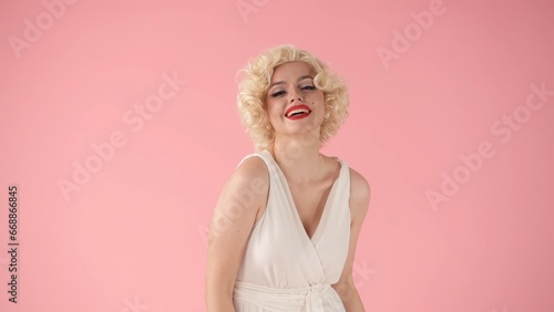 Portrait young woman in wig, white dress and with red lipstick on lips in studio on pink background. Woman looking like Marilyn Monroe in studio on pink background.