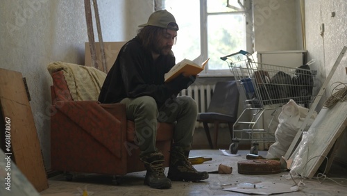 Homeless poor man sitting in a room of an abandoned building filled with his meager belongings. He is reading a book, flipping through the pages.