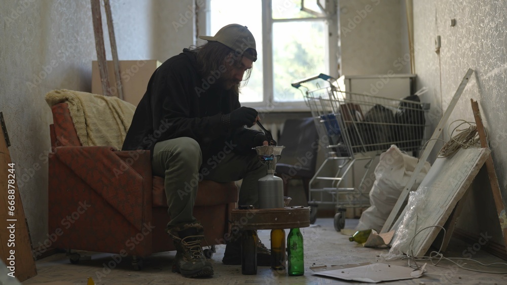 Homeless poor man sitting in a room of an abandoned building filled with his meager belongings. He is heating his food on a gas burner. Homeless poor man sitting in a room of an abandoned building filled with his meager belongings. He is heating his food on a gas burner.