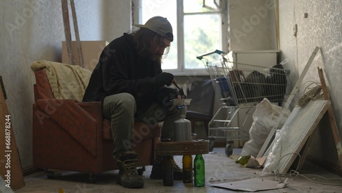 Homeless poor man sitting in a room of an abandoned building filled with his meager belongings. He is heating his food on a gas burner.