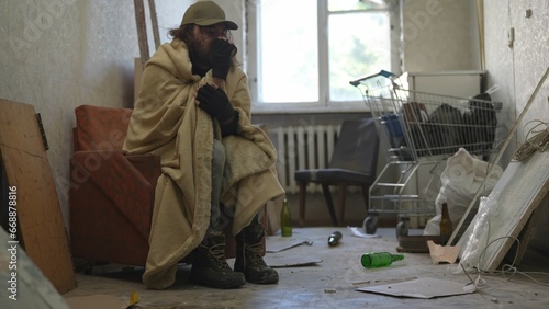 Homeless poor man sitting in a room of an abandoned building. He is trying to keep warm under a blanket, drinking warm beverage from a cup.