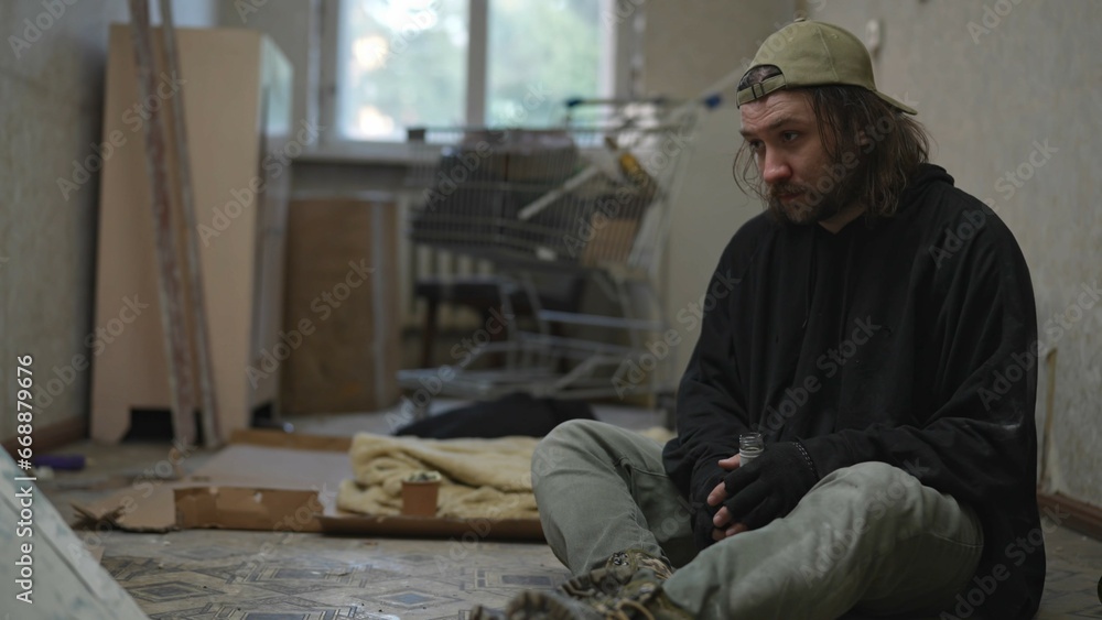 Homeless poor man sitting in a room of an abandoned building filled with his meager belongings. He is holding a bottle of alcohol in his hand, looking frustrated. Homeless poor man sitting in a room of an abandoned building filled with his meager belongings. He is holding a bottle of alcohol in his hand, looking frustrated.