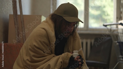 Homeless poor man covered in a blanket sitting in a room of an abandoned building filled with his meager belongings. He is holding a bottle of pills. Medium shot.