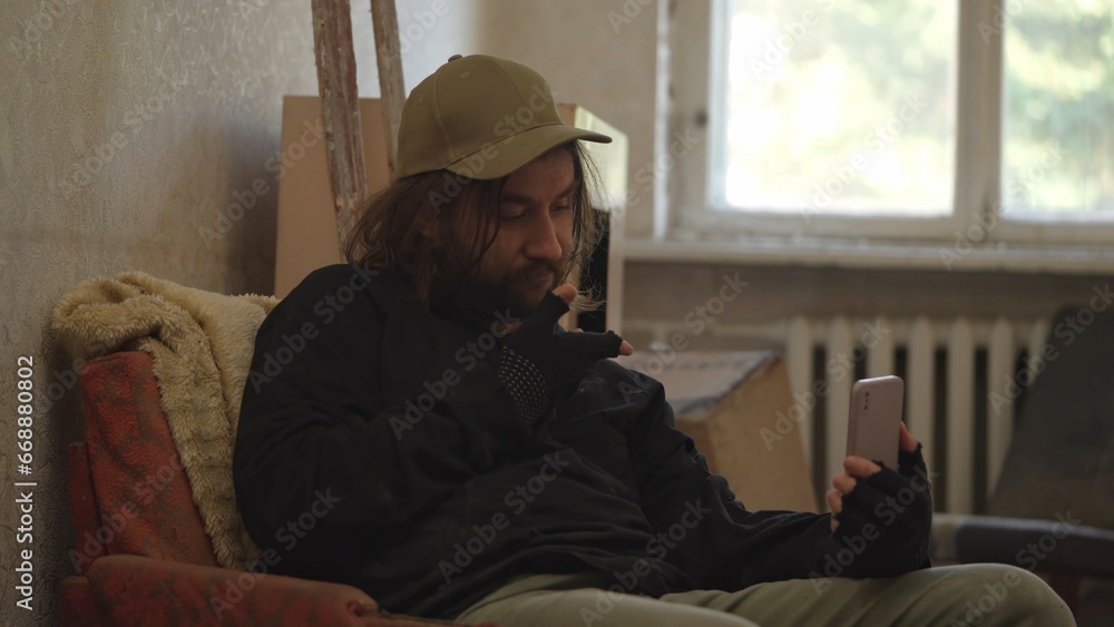 Homeless poor man sitting in a room of an abandoned building filled with his meager belongings. He is having a videocall, showing the place he is in. Medium shot. Homeless poor man sitting in a room of an abandoned building filled with his meager belongings. He is having a videocall, showing the place he is in. Medium shot.