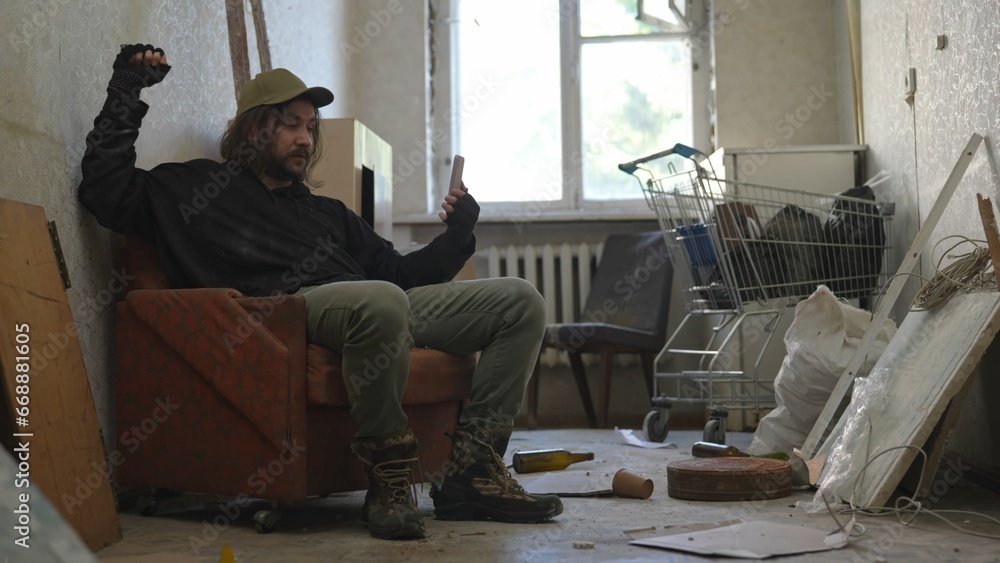 Homeless poor man sitting in a room of an abandoned building filled with his meager belongings. He is having a videocall, showing the place he is in.