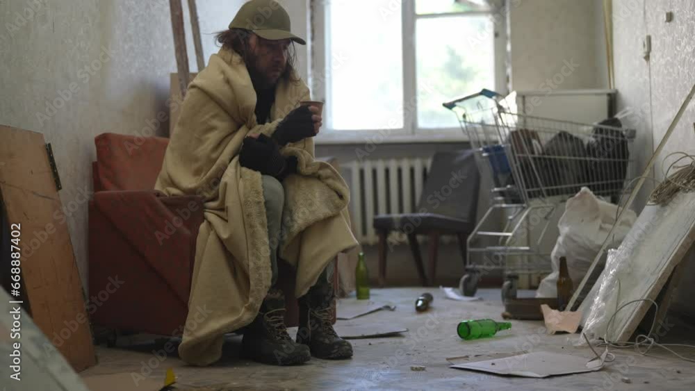 Homeless poor man sitting in a room of an abandoned building. He is trying to keep warm under a blanket, drinking warm beverage from a cup. Homeless poor man sitting in a room of an abandoned building. He is trying to keep warm under a blanket, drinking warm beverage from a cup.