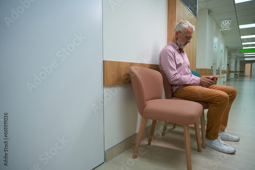 Elderly man sits with a mobile phone in hospital corridor