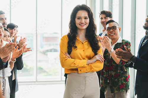 Successful businesswoman looking at camera with confidence while standing rounded with her coworker clapping hands. Diverse team celebrates promotion of new beautiful manager. Intellectual.