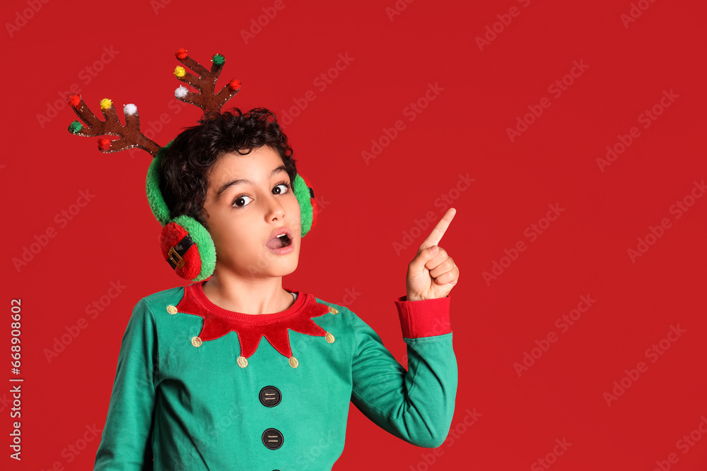 Cute little boy in elf costume with deer horns pointing at something on red background Cute little boy in elf costume with deer horns pointing at something on red background