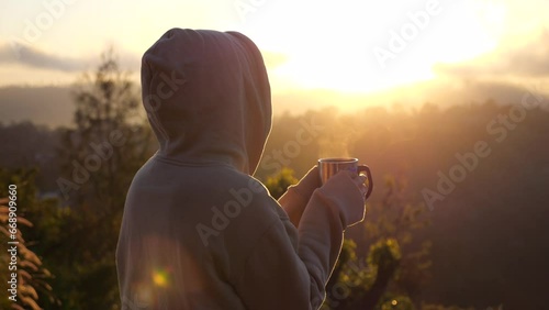 Hiker woman standing on cliff against background of sunrise, holding coffee mug with steam Close-up of hiker in cozy hoodie in cold early morning watching sunrise Travel on camping van to meet sunrise