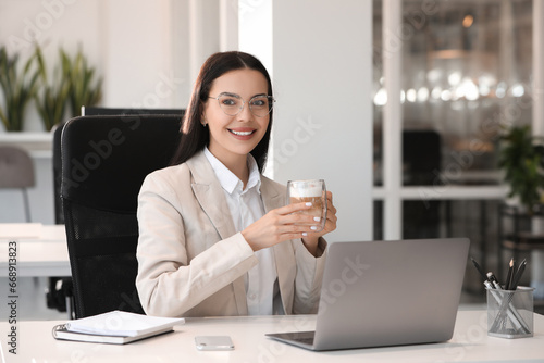 Happy woman with cup of coffee using modern laptop at white desk in office