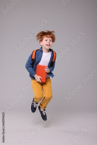 Happy schoolboy with backpack and book jumping on grey background