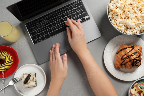 Bad eating habits. Woman using laptop surrounded by different snacks at grey table, top view
