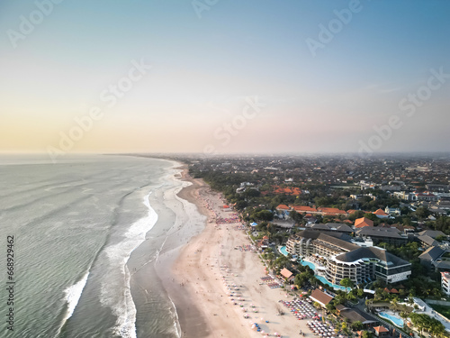 Aerial view of Beautiful sunset on the double six beach with Hotel, resort, cafe buildings, beach umbrella and calm ocean wave in Bali. Tourists are surfing, swimming and walking along the beach.