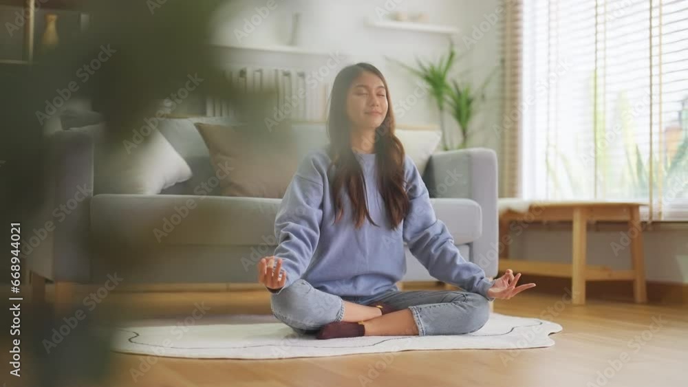 Happy young Asian woman practicing yoga and meditation at home sitting on floor in living room in lotus position and relaxing with closed eyes. Mindful meditation and wellbeing concept