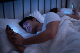 Asian insomnia couple boy and girl addicted playing social media and texting message to friend with blue light from mobile screen when turned their backs sleep in bed at the dark night bedroom