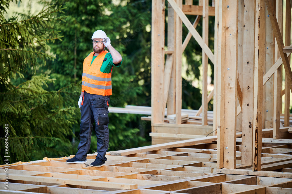 Carpenter constructing wooden frame two-story house near the forest. Bearded man with glasses dressed in protective helmet and orange safety vest. Concept of ecological modern construction. Carpenter constructing wooden frame two-story house near the forest. Bearded man with glasses dressed in protective helmet and orange safety vest. Concept of ecological modern construction.
