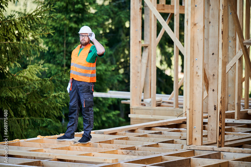Carpenter constructing wooden frame two-story house near the forest. Bearded man with glasses dressed in protective helmet and orange safety vest. Concept of ecological modern construction.