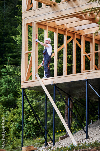 Carpenter constructing two-story wooden frame house in close proximity to woods. Bearded man  dressed in jumpsuit and protective helmet  assesses the wall s levelness with the aid of a spirit level.
