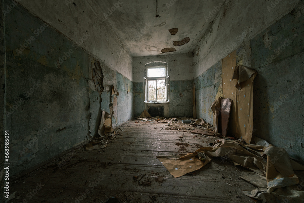 Obraz premium Interior of abandoned room with peeling painted walls damaged wooden furniture garbage on dirty floor with crumbling ceiling showing iron rods with glass window in daylight