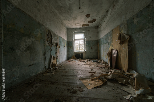 Interior of abandoned room with peeling painted walls damaged wooden furniture garbage on dirty floor with crumbling ceiling showing iron rods with glass window in daylight