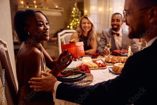 Happy black woman receives Christmas present during dinner party.