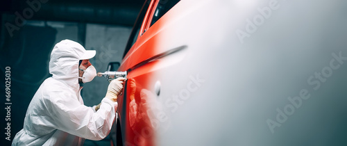 A car painter in a white protective suit paints a car with red spray paint. Copy space. Banner.