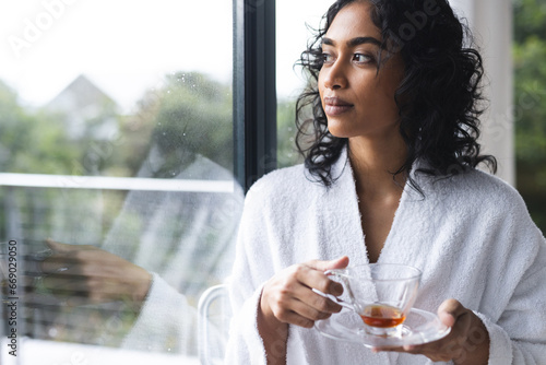 Happy biracial woman in bathrobe holding cup of tea and looking out window in sunny room at home photo